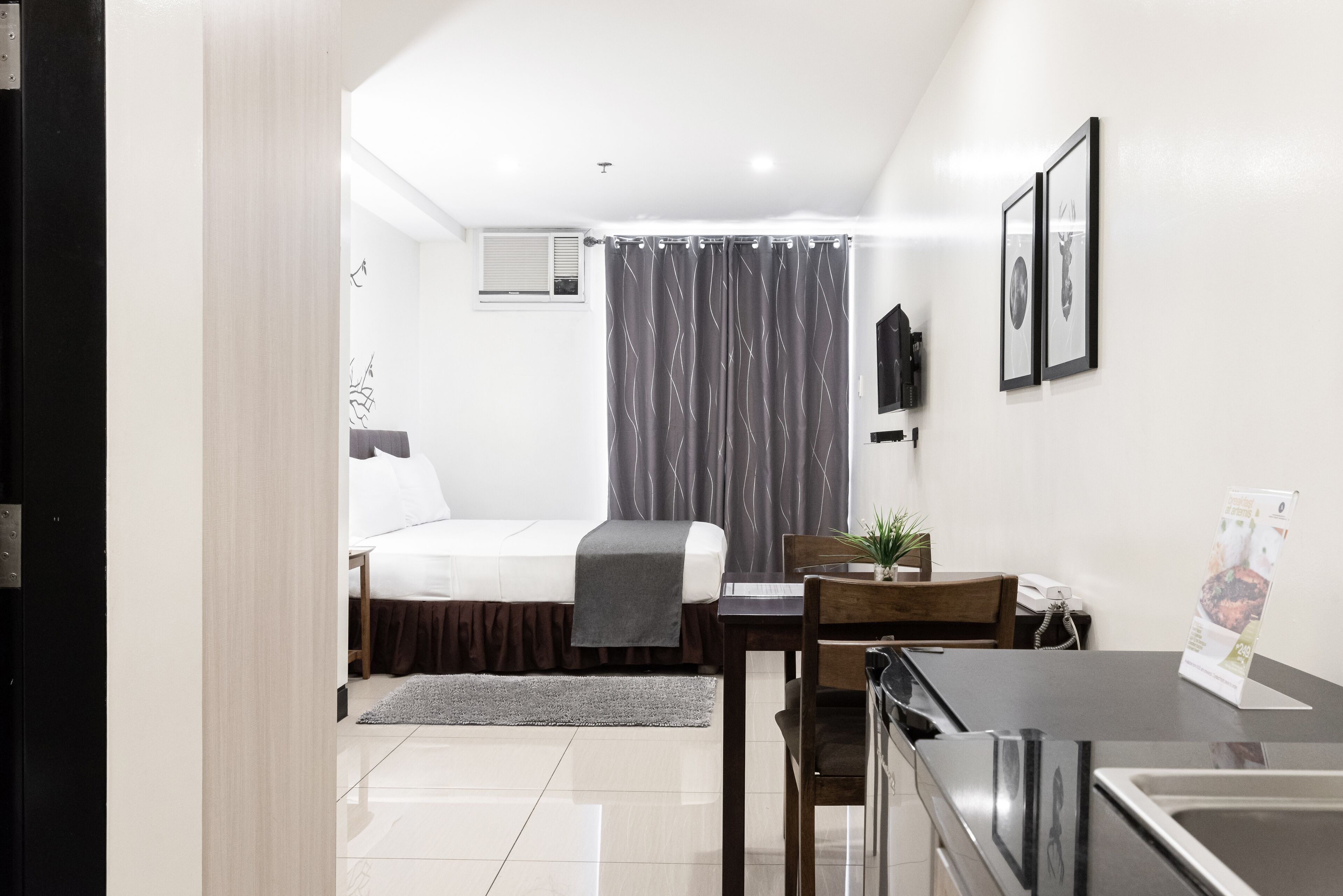 Une chambre d’hôtel avec un lit, un bureau, une chaise, une petite table, une plante, un cadre photo et une fenêtre avec des rideaux.