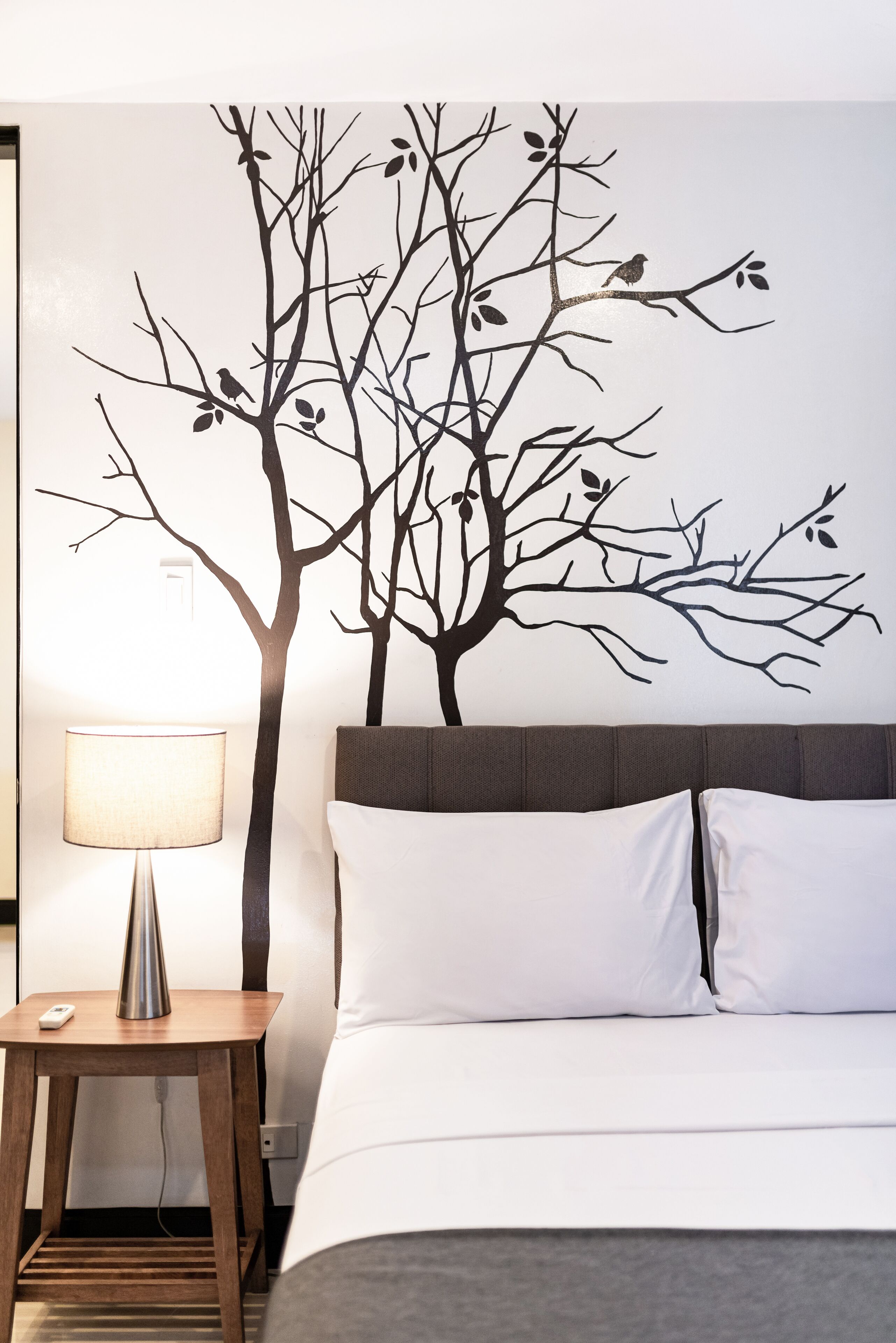 A bedroom with a bed, a nightstand, a lamp, and a wall decal of a tree with birds.