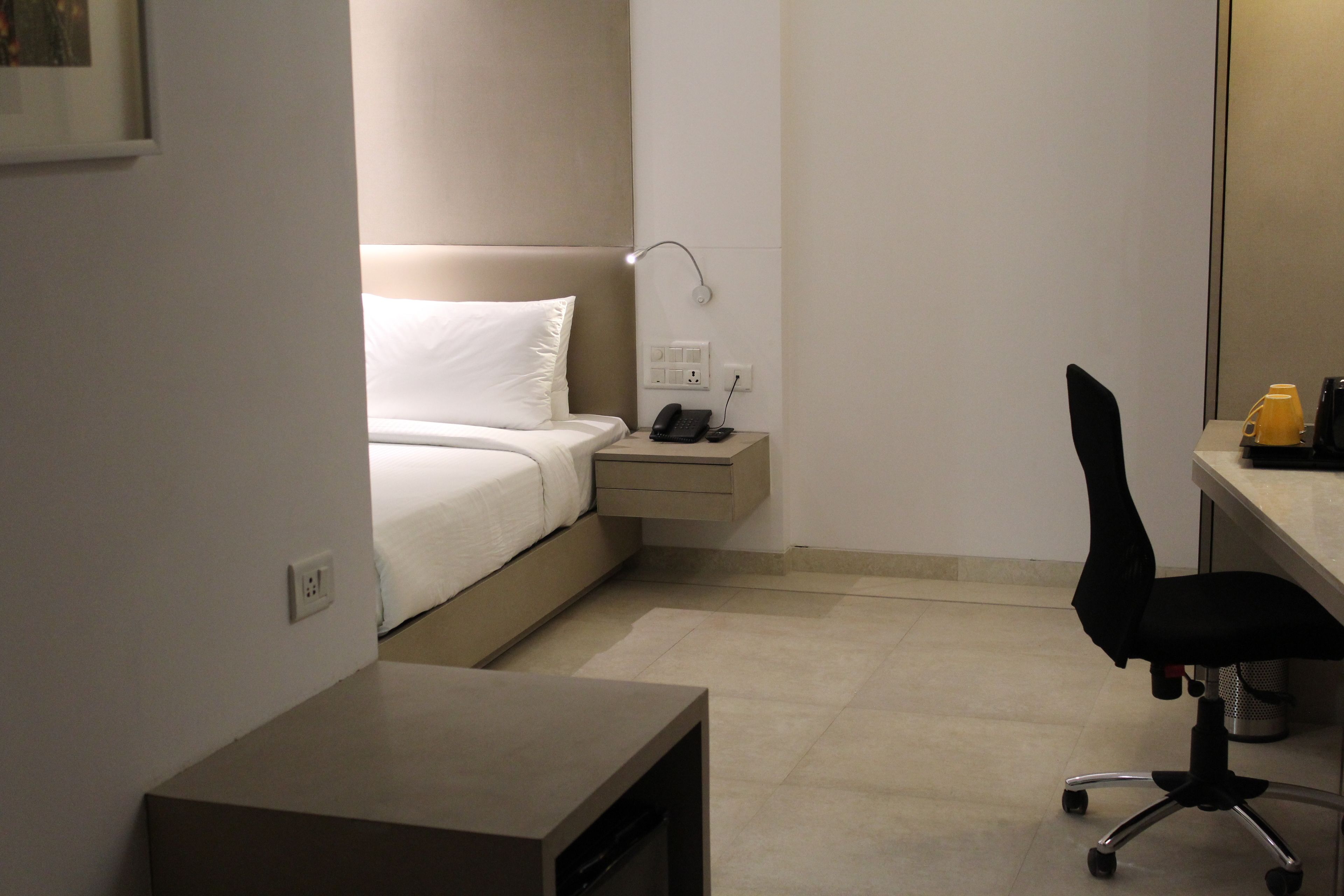 A hotel room with a bed, bedside table, lamp, desk, chair, and a small trash can.