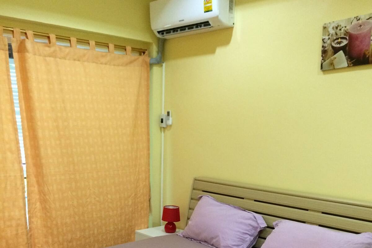 Un lit simple avec une tête de lit en bois, une table de chevet avec une lampe rouge et un appareil de climatisation fixé au mur.