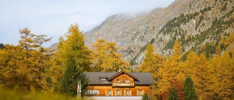 A wooden chalet with a balcony, surrounded by autumn-colored trees and mountains in the background.
