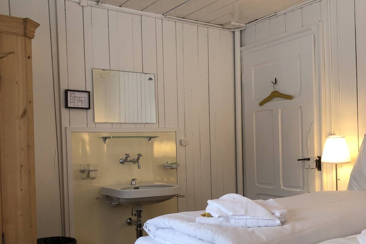 A neatly made bed with white linens, a bedside table with a lamp, a wooden cabinet, and a bathroom with a sink and mirror.