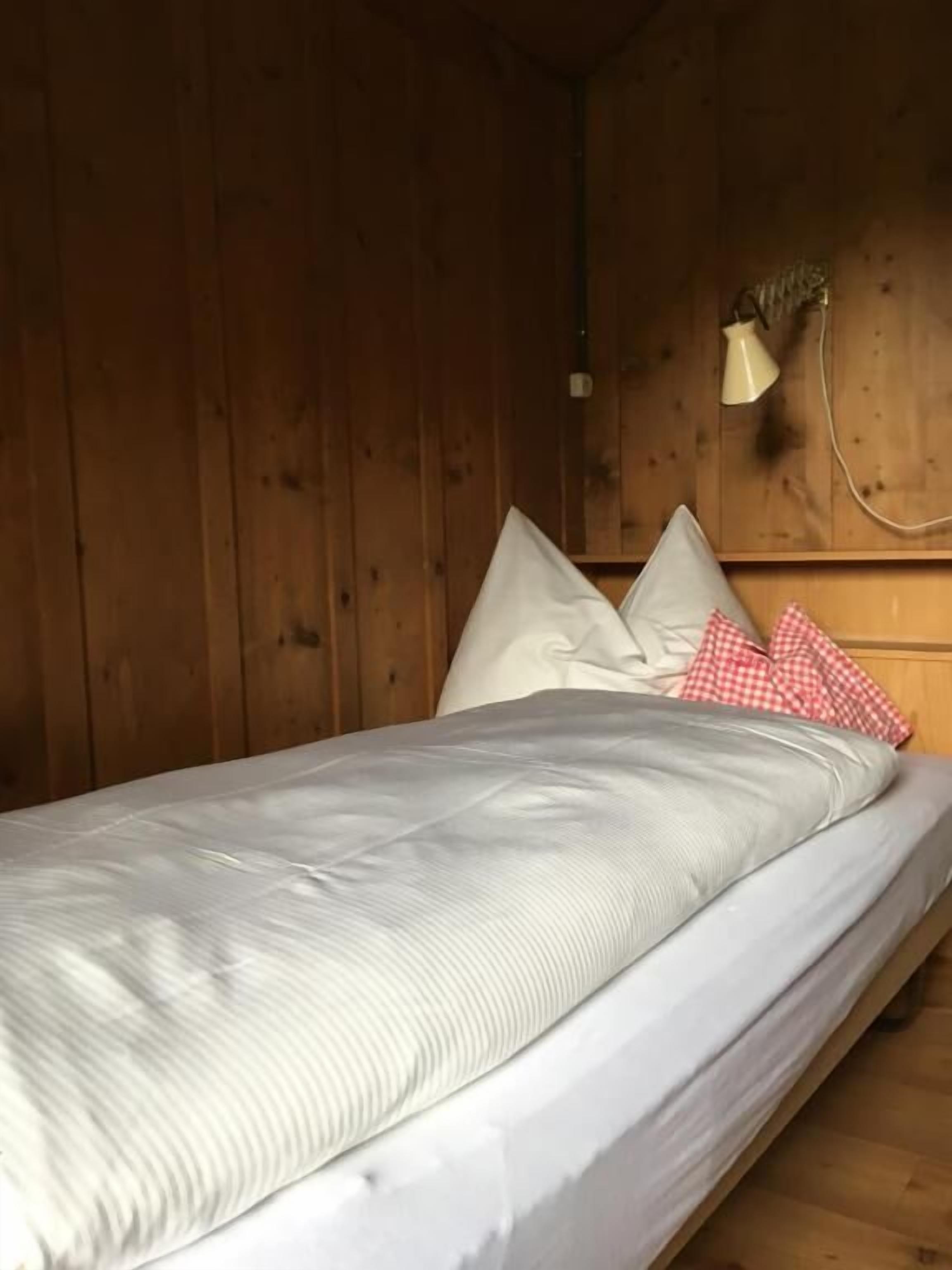Un lit simple avec un matelas et des oreillers blancs, une tête de lit en bois et une lampe fixée au mur.