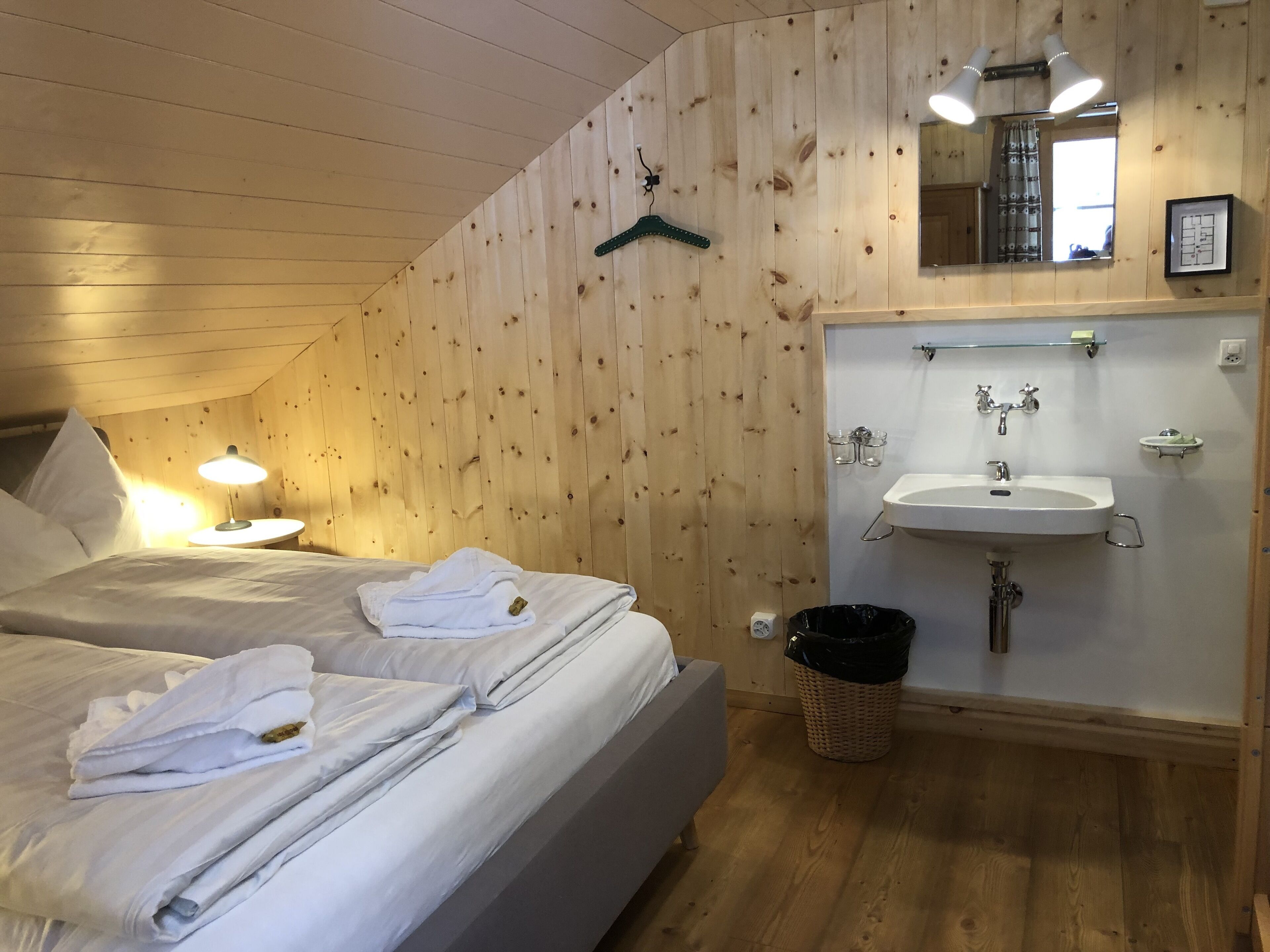 Un dormitorio con dos camas, una mesita de noche con lámpara, un muro de madera, un perchero y un baño con lavamanos y espejo, además de un cubo de basura.