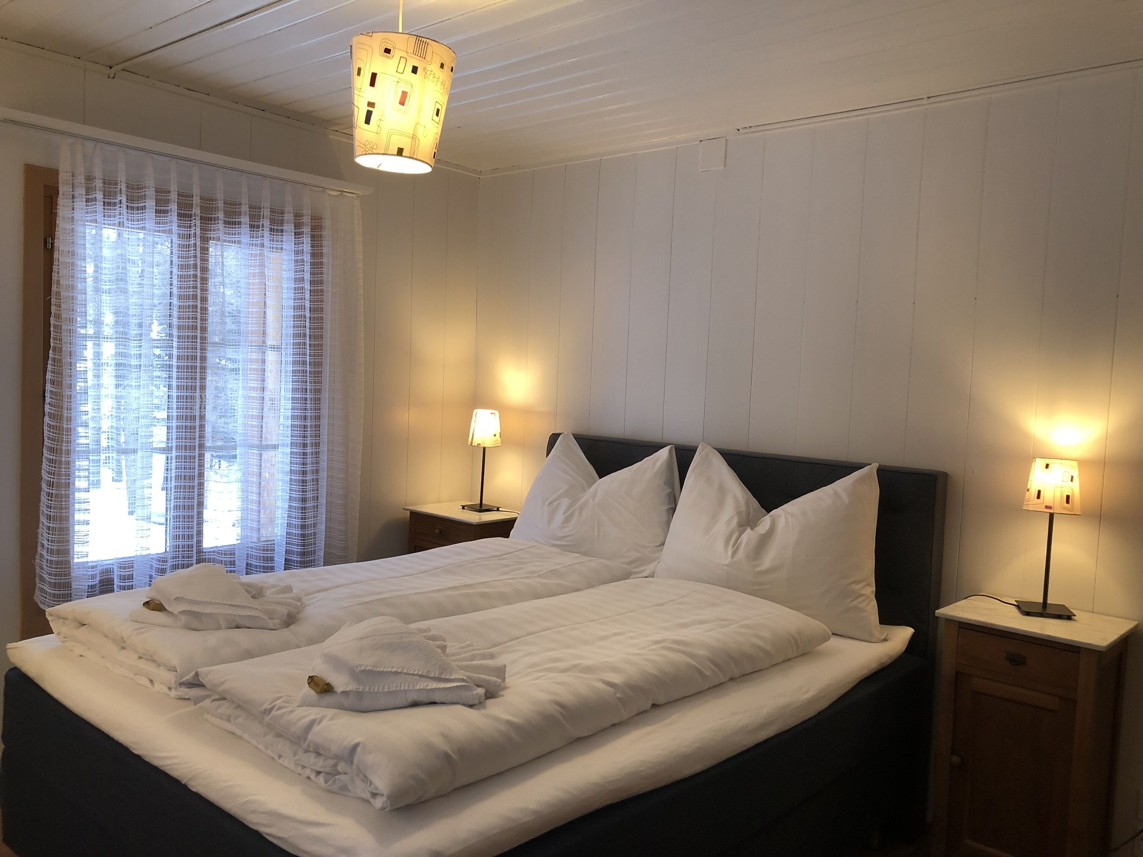 Una cama bien hecha con sábanas blancas, dos mesitas de noche con lámparas y una ventana con cortinas traslúcidas.