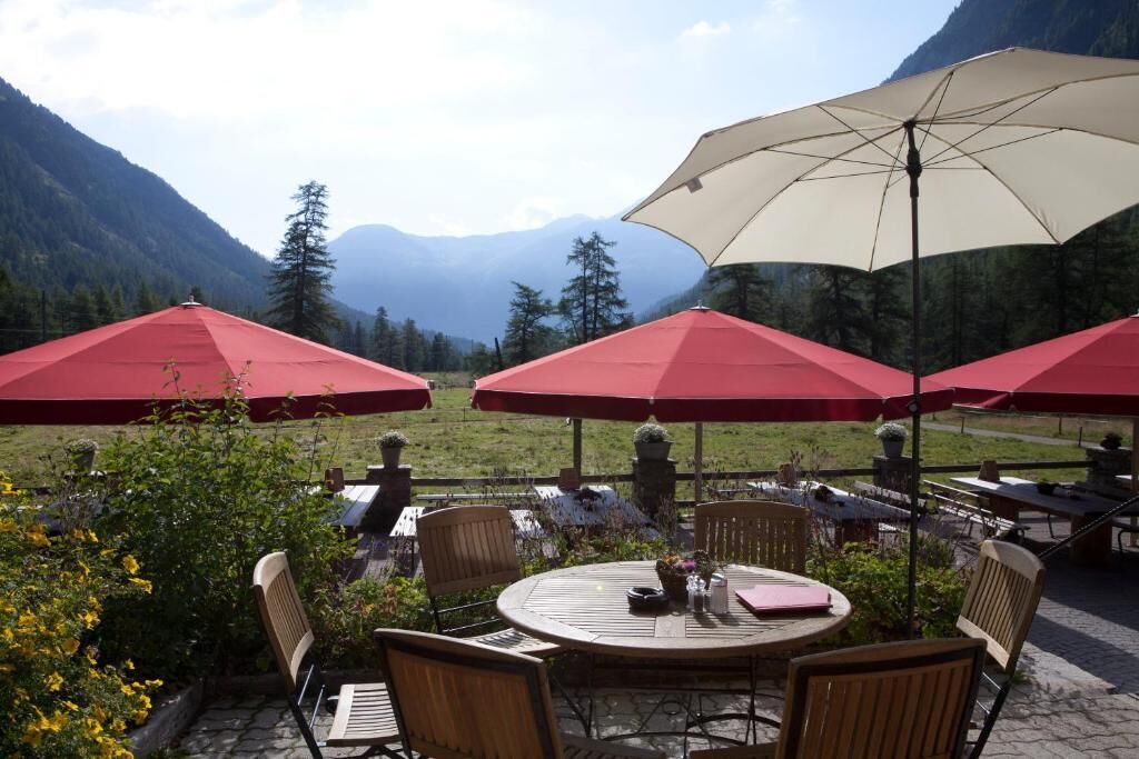 Ein Sitzbereich im Freien mit Tischen und Stühlen, umgeben von roten Sonnenschirmen, vor einer Bergkulisse.