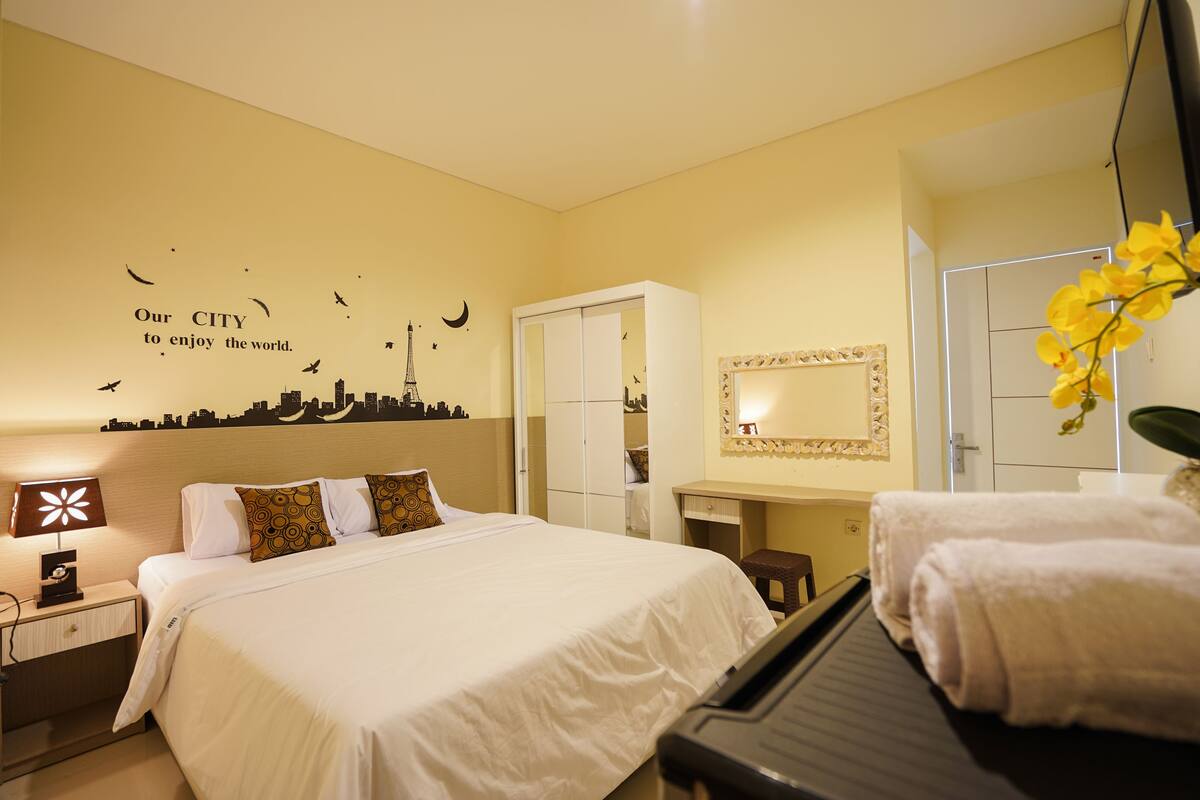 A hotel room with a bed, a desk, a mirror, and a piano.