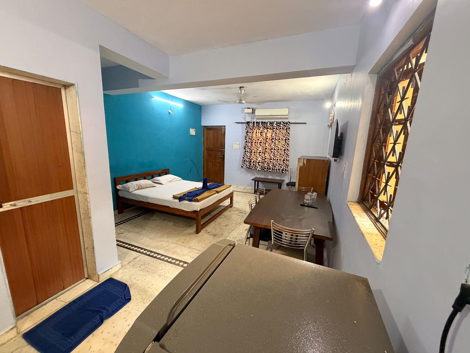 Quarto compacto com cama, mesa de jantar e TV fixada na parede.