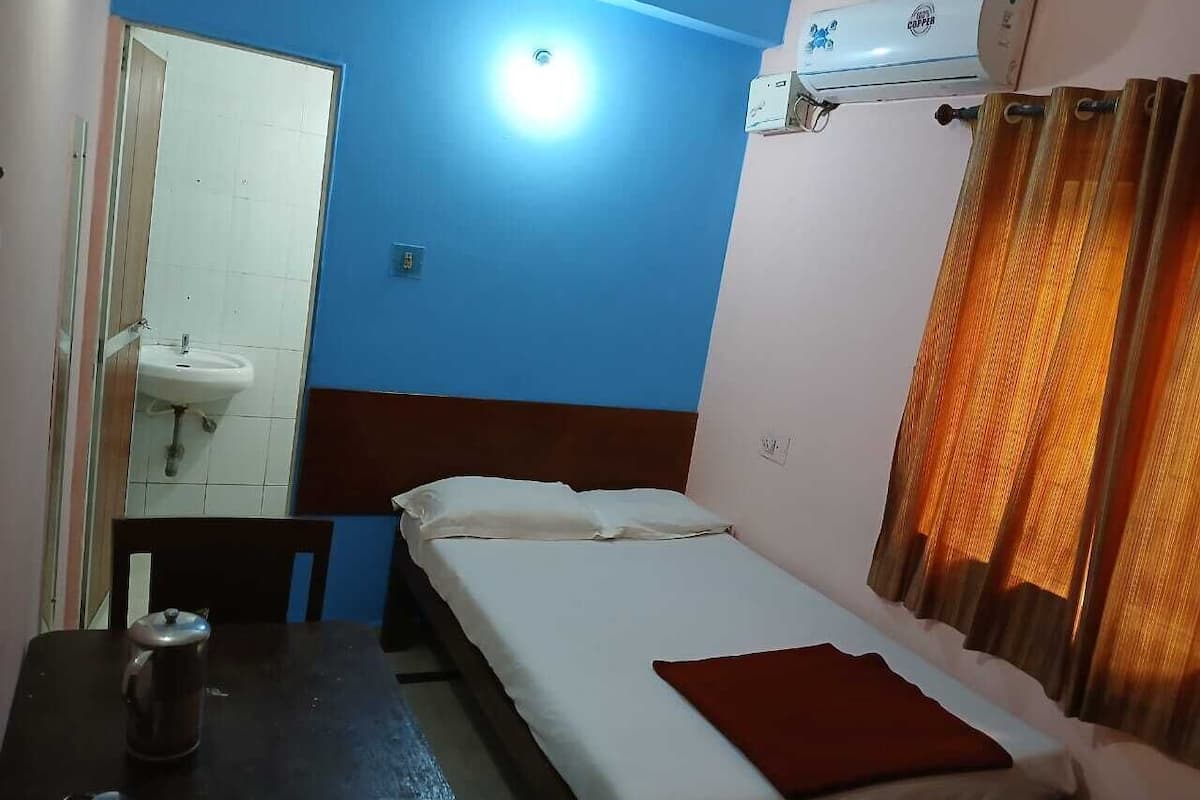 A small, single-bed room with a white sink, a table with a kettle, and a window with orange curtains.