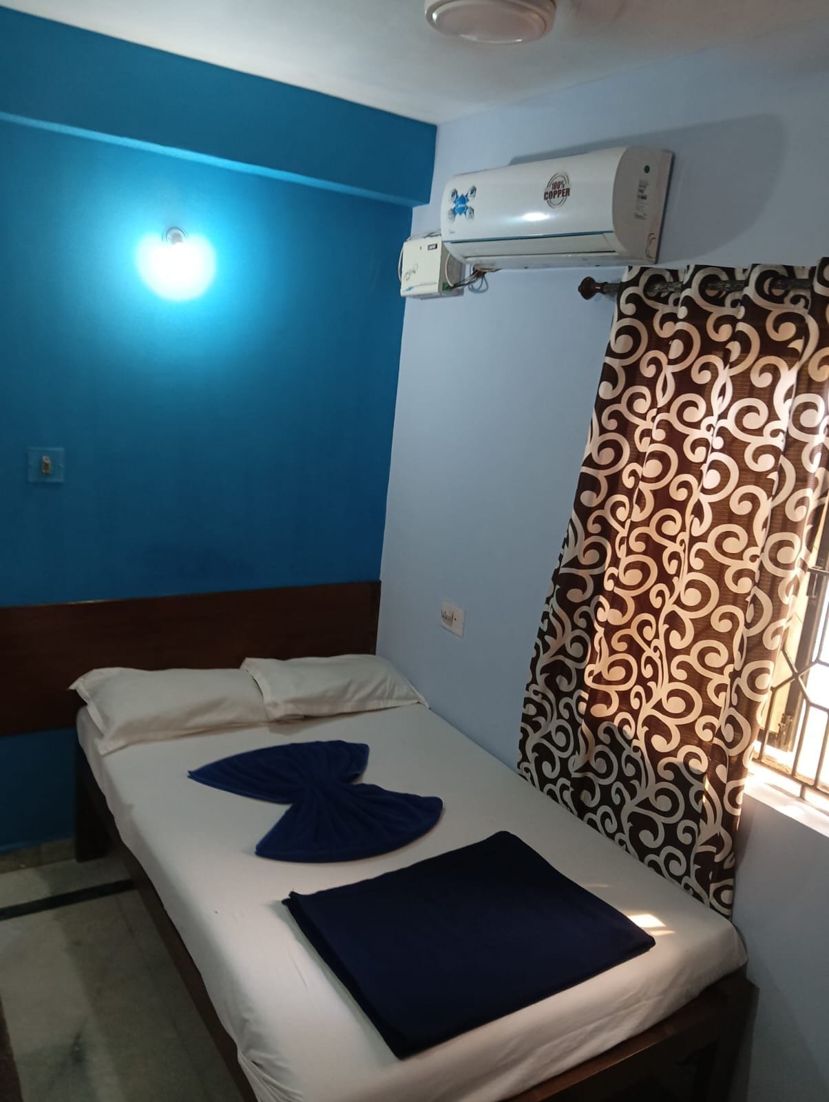 A single bed with a white mattress and blue pillows, a wall-mounted air conditioner, and a window with patterned curtains.