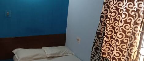 A single bed with a white mattress and blue pillows, a wall-mounted air conditioner, and a window with patterned curtains.