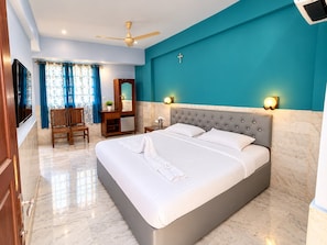 A hotel room with a large bed, a ceiling fan, a TV, a desk with a chair, and a cross on the wall. - Blue Marine Resort (Calangute)
