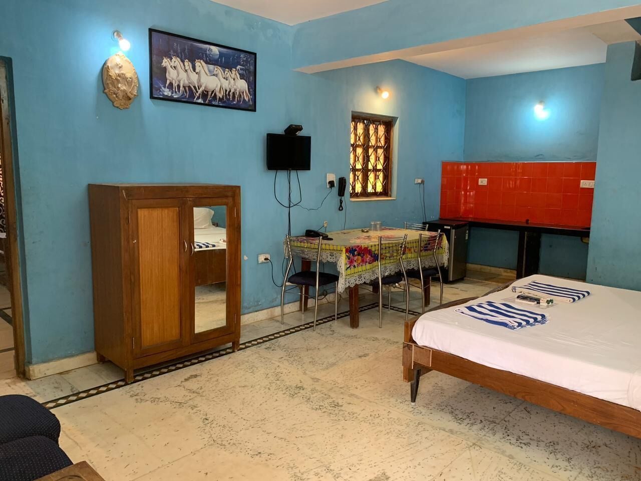 Un dormitorio con una cama, un armario de madera, una mesa con mantel floreado y un televisor montado en la pared.
