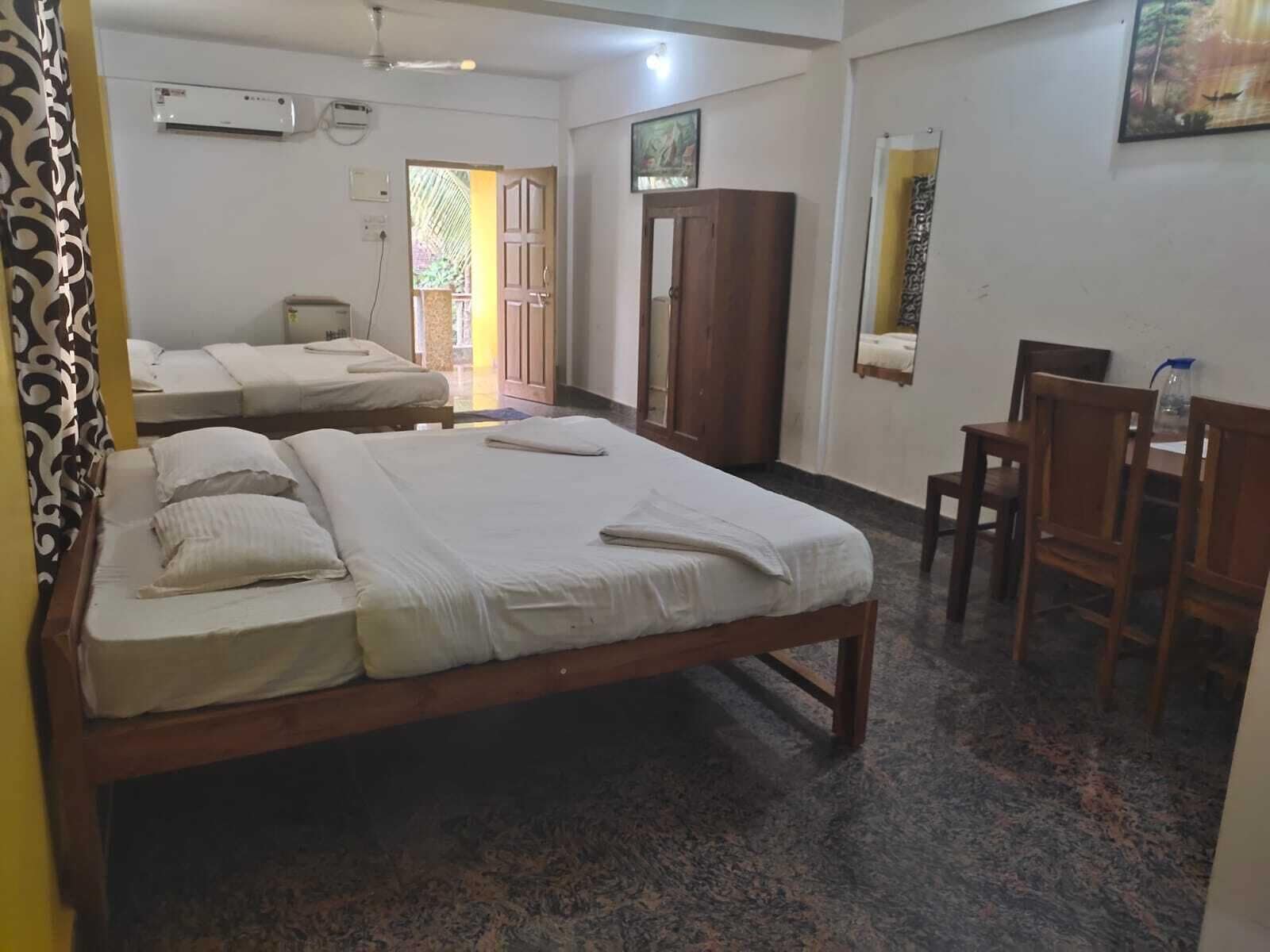 A hotel room with two single beds, a wooden wardrobe, a desk with chairs, and an air conditioning unit on the wall.