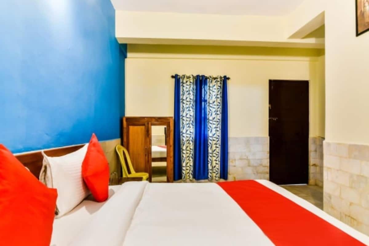 A hotel room with a bed, red and white bedding, a wooden chair, a door, and blue patterned curtains.