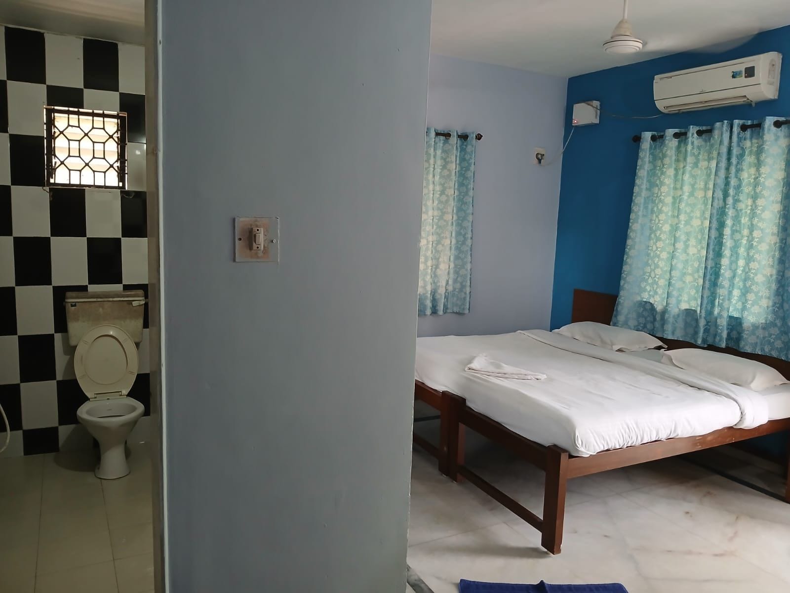 A room with a bed, a window with curtains, a wall-mounted air conditioner, and a checkered wall pattern.