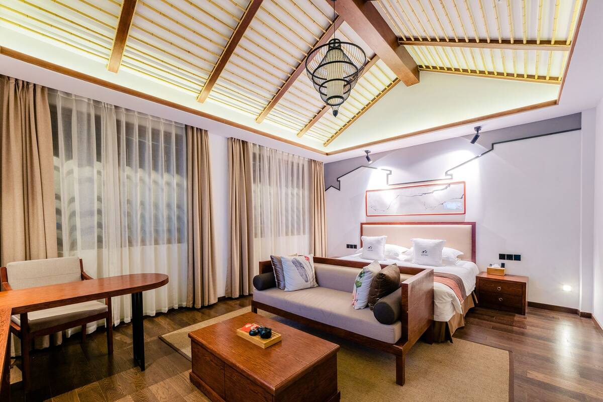 A modern hotel room with a wooden ceiling, a sofa bed with pillows, a wooden coffee table, and a bedside table.