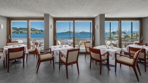 A dining area with tables and chairs, large windows with a view of the sea and hills. - Alkyon Hotel (Skiathos)