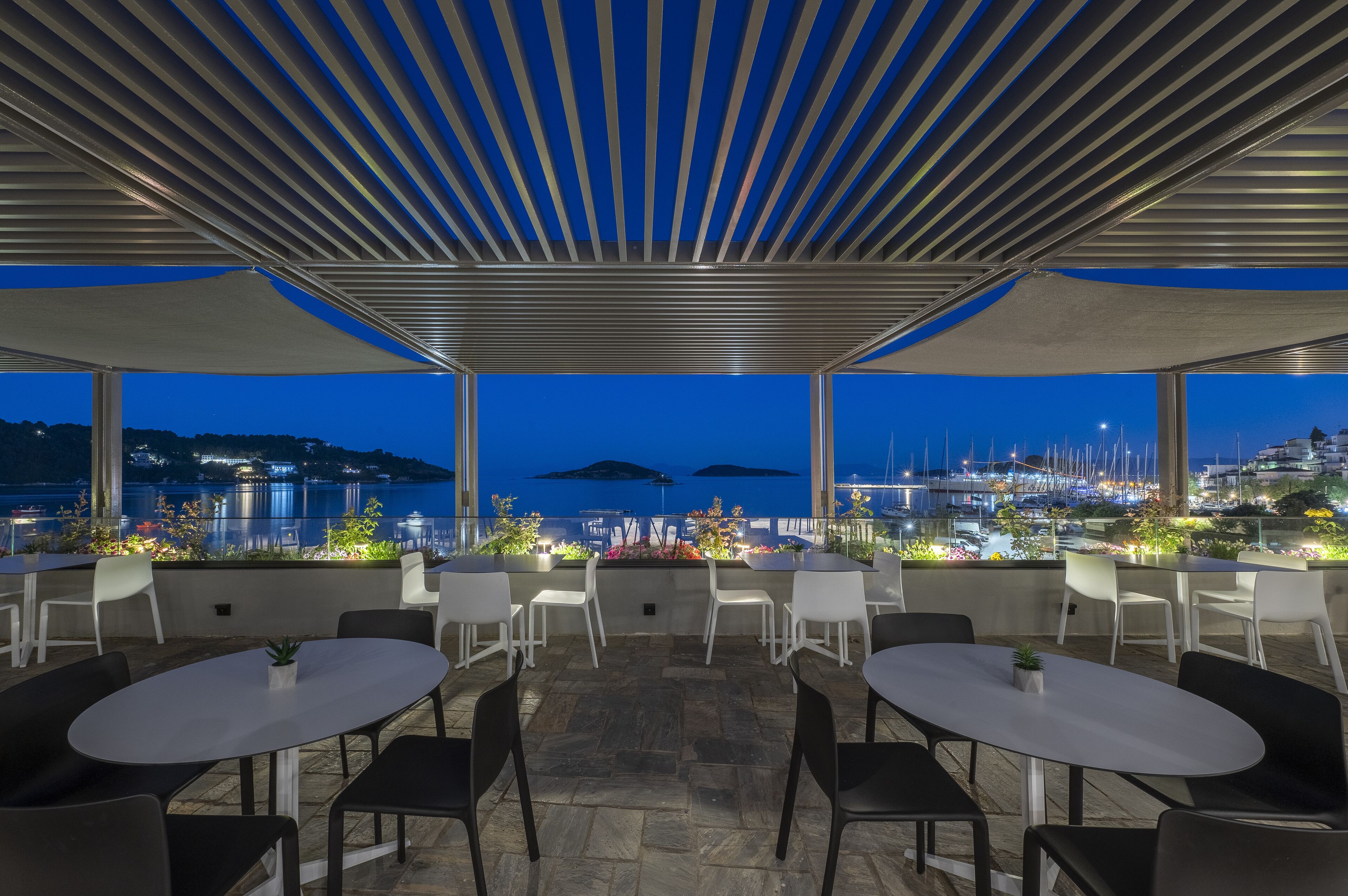 Terraza al aire libre con mesas y sillas, con vistas a un puerto deportivo y al paisaje urbano al anochecer.