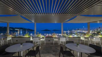 Terraza al aire libre con mesas y sillas, con vistas a un puerto deportivo y al paisaje urbano al anochecer.