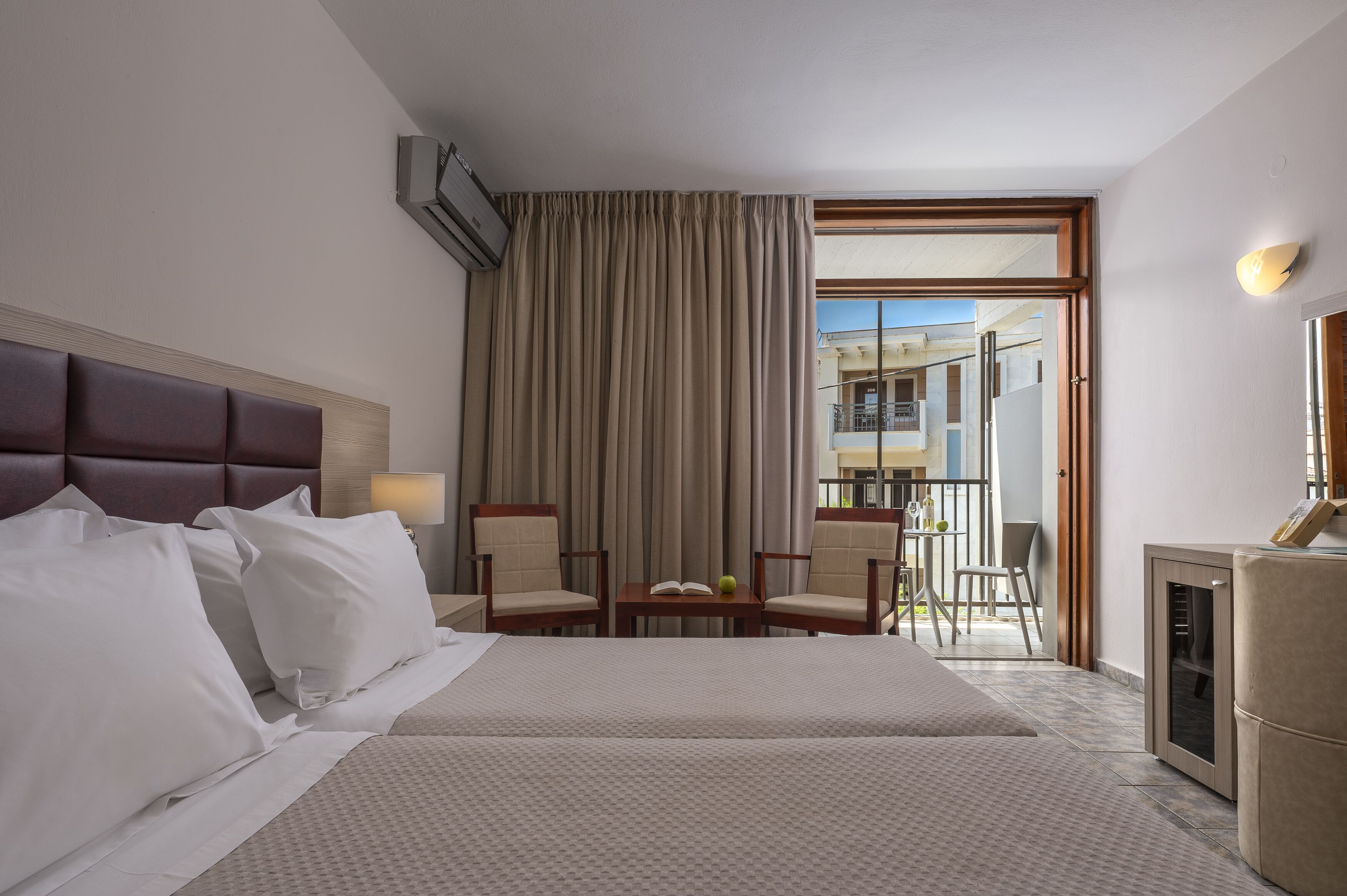 A hotel room with a bed, two chairs, a small table with a book and fruit, and a view of the outside balcony.