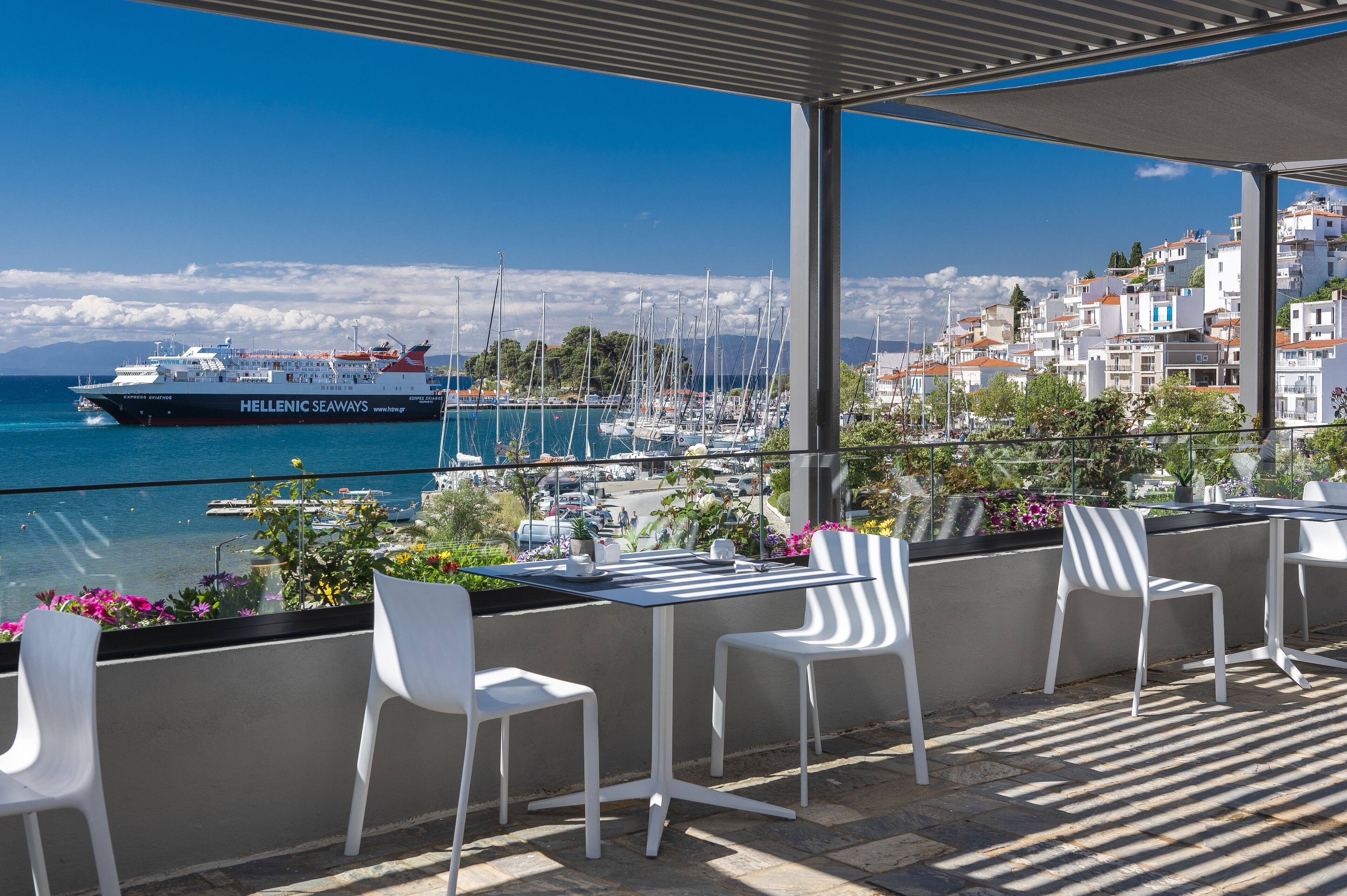 Eine Terrasse mit weißen Stühlen und einem Tisch mit Blick auf eine Marina, in der Segelboote und ein großes Schiff namens "HELLENIC SEAWAYS" liegen.