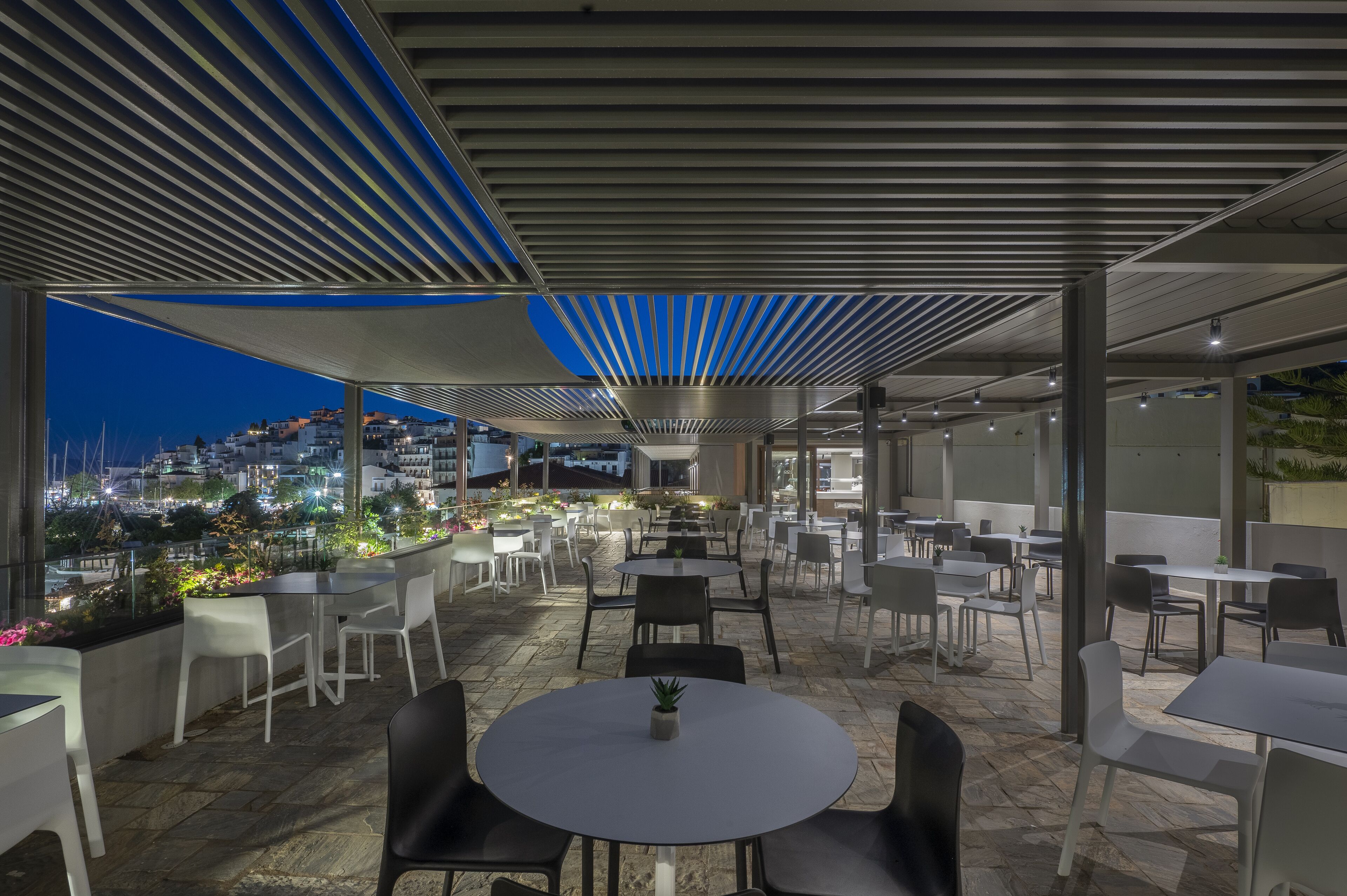 An outdoor dining area with tables and chairs, a view of the cityscape, and a covered section with a blue-lit ceiling.