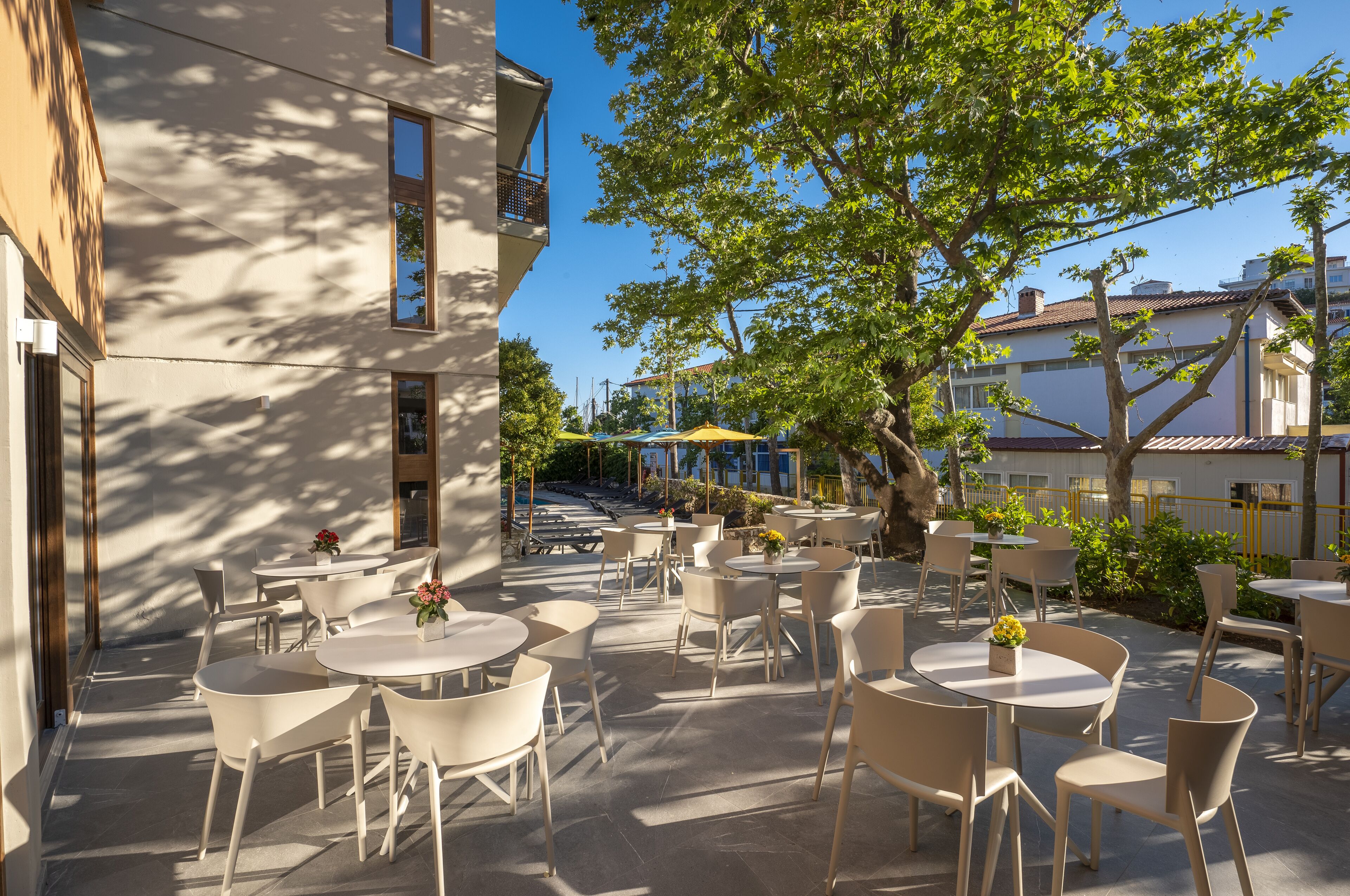 An outdoor cafe area with round tables and chairs, surrounded by buildings and trees.