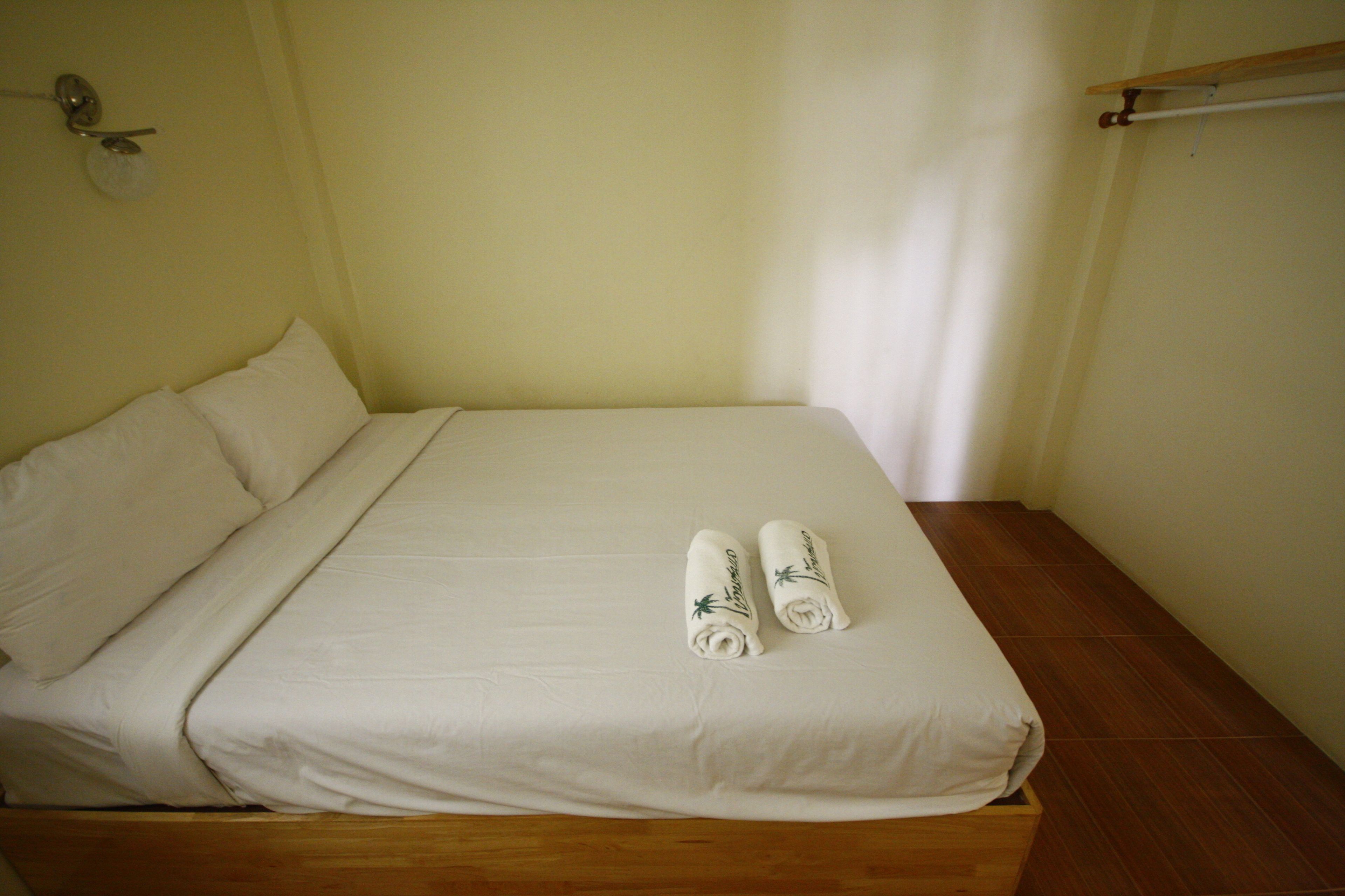 Un lit simple avec un cadre en bois et du linge de lit blanc, deux serviettes enroulées ornées de motifs de palmiers, et une étagère en bois avec un luminaire placé au-dessus.