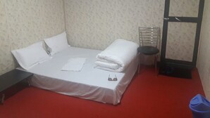 Ein ordentlich bezogenes Bett mit weißen Leinen und Kissen, eine Sonnenbrille auf dem Bett, ein kleiner runder Tisch mit einem Stuhl und eine blaue Mülltonne.