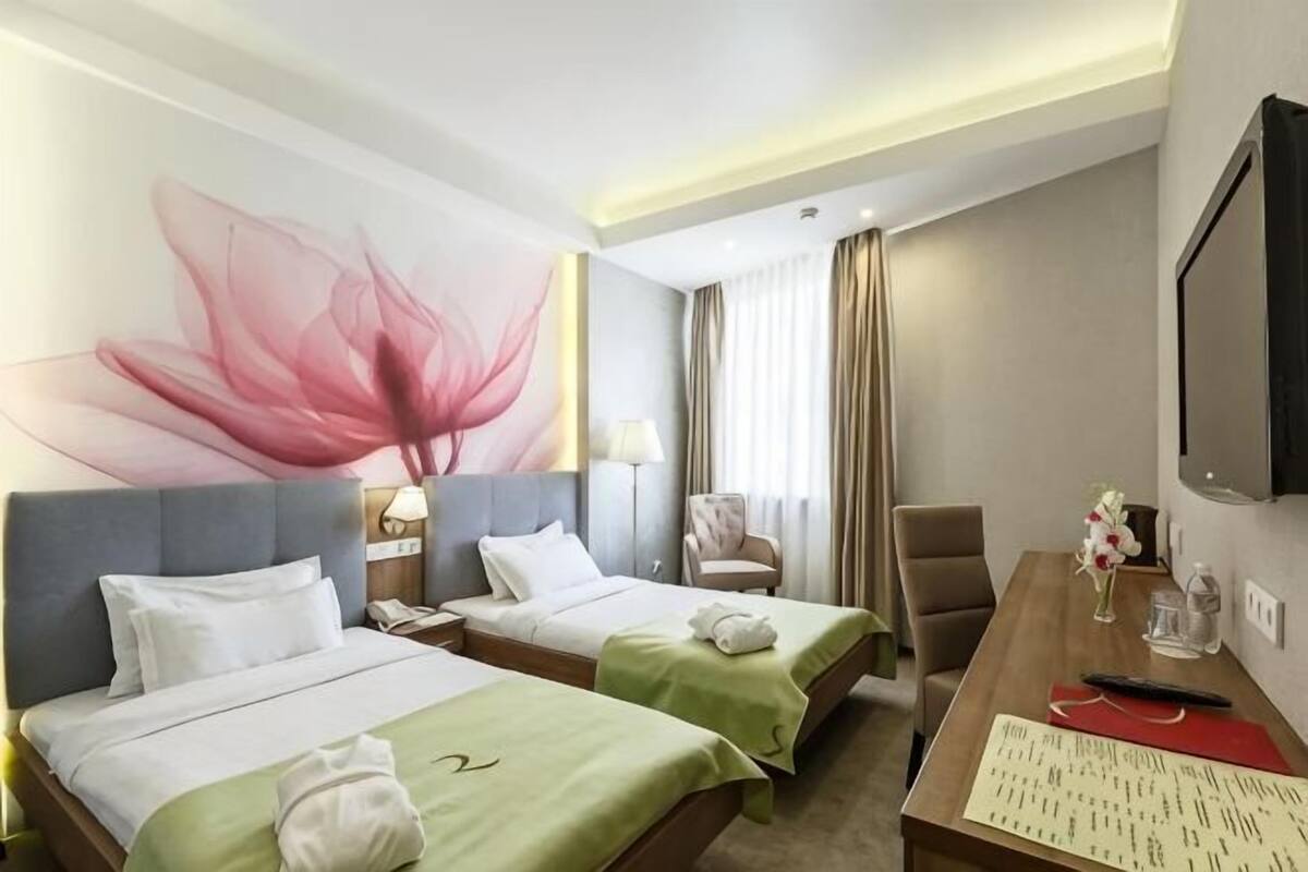 A hotel room with two beds, a desk with a keyboard, a TV, and a large floral artwork on the wall.