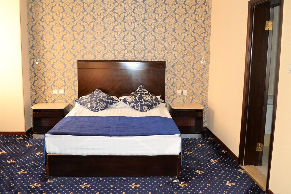 A neatly made bed with a blue and white bedspread, two matching pillows, and a wooden headboard. There are two bedside tables with lamps, and the room has patterned wallpaper and carpet.