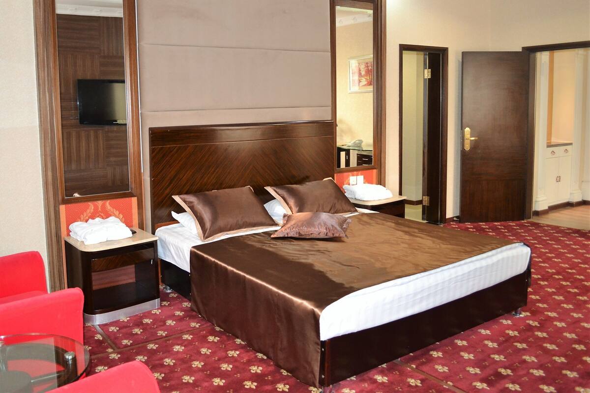 A hotel room with a large bed, a bedside table, a red chair, a glass table, and a mirror.