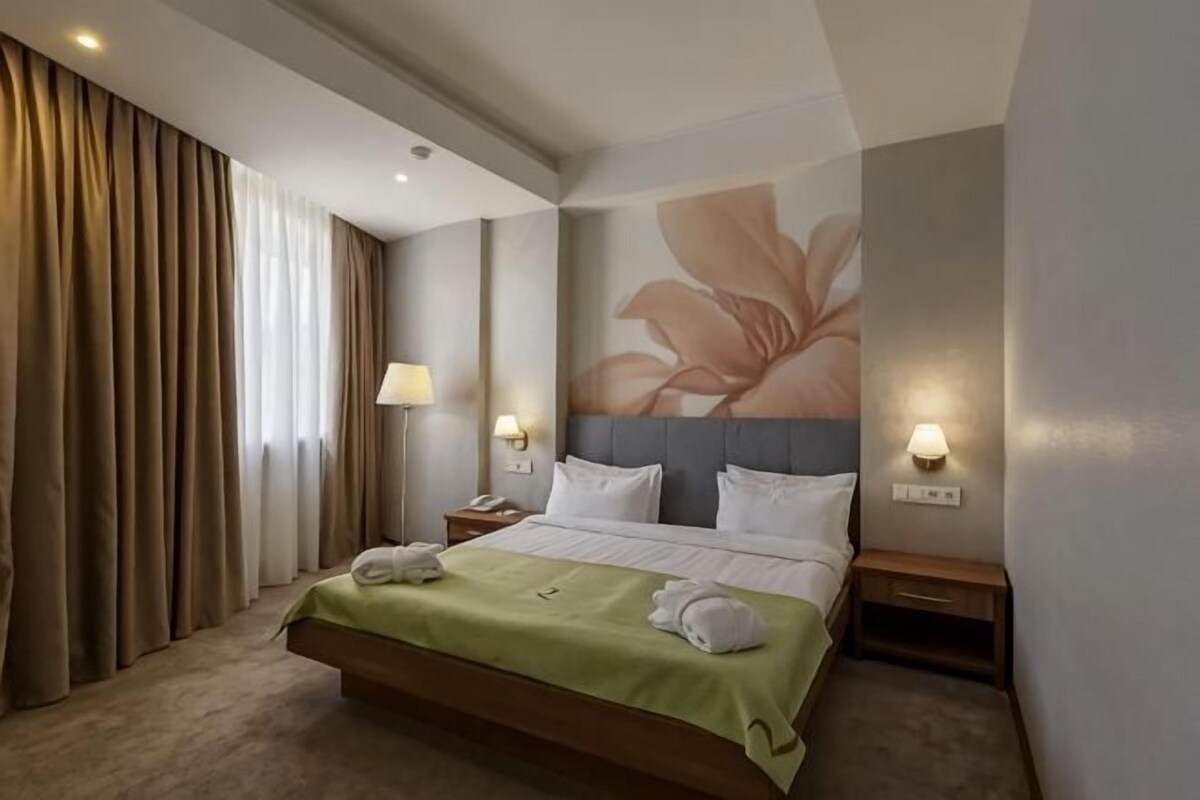 Habitació d'hotel amb un llit, dues taules de nit, una tauleta auxiliar, una làmpada de paret i un quadre floral gran.