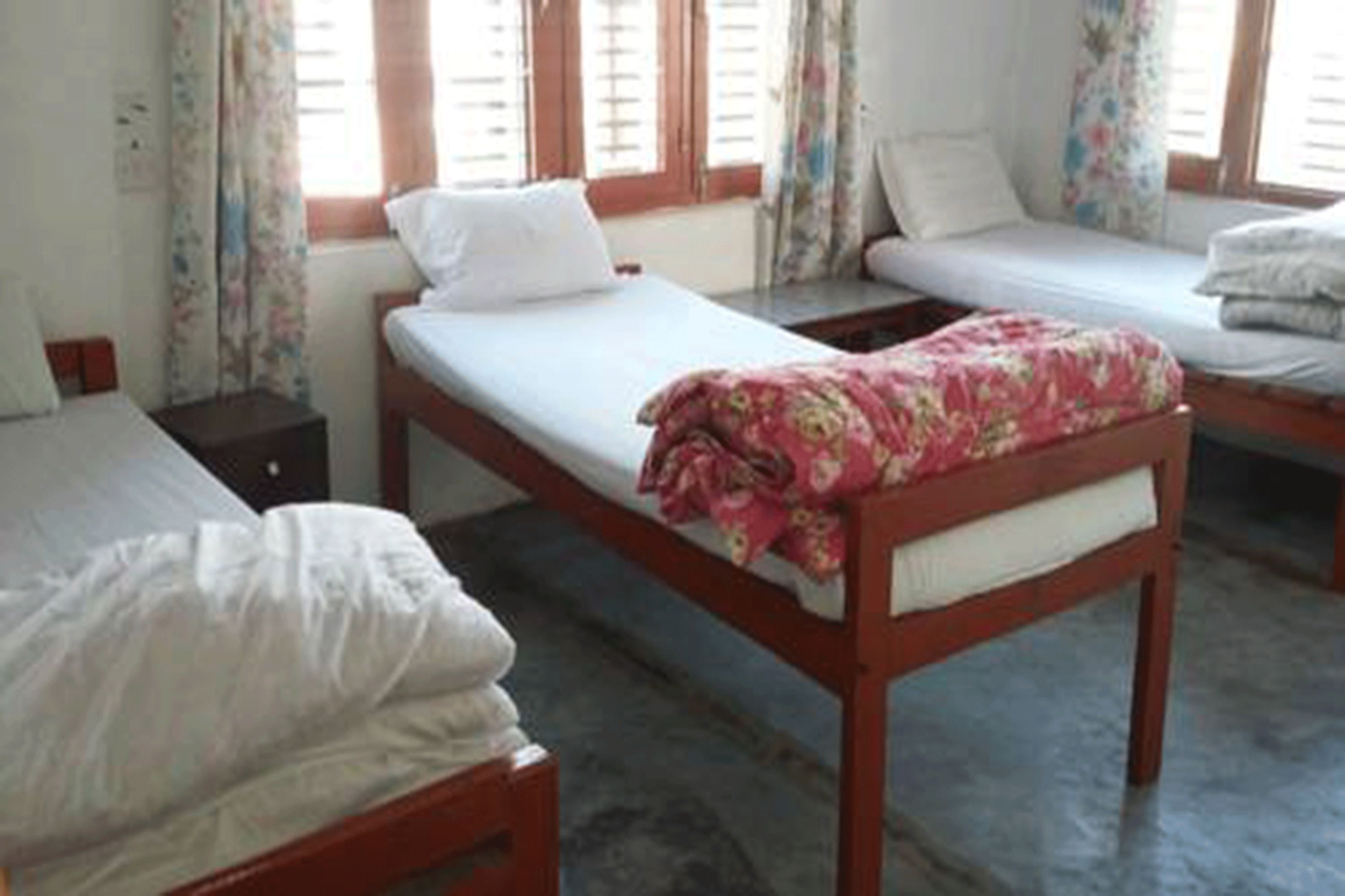 A room with three single beds, each with a pillow and a blanket. There are curtains on the windows, and the room has a tiled floor.