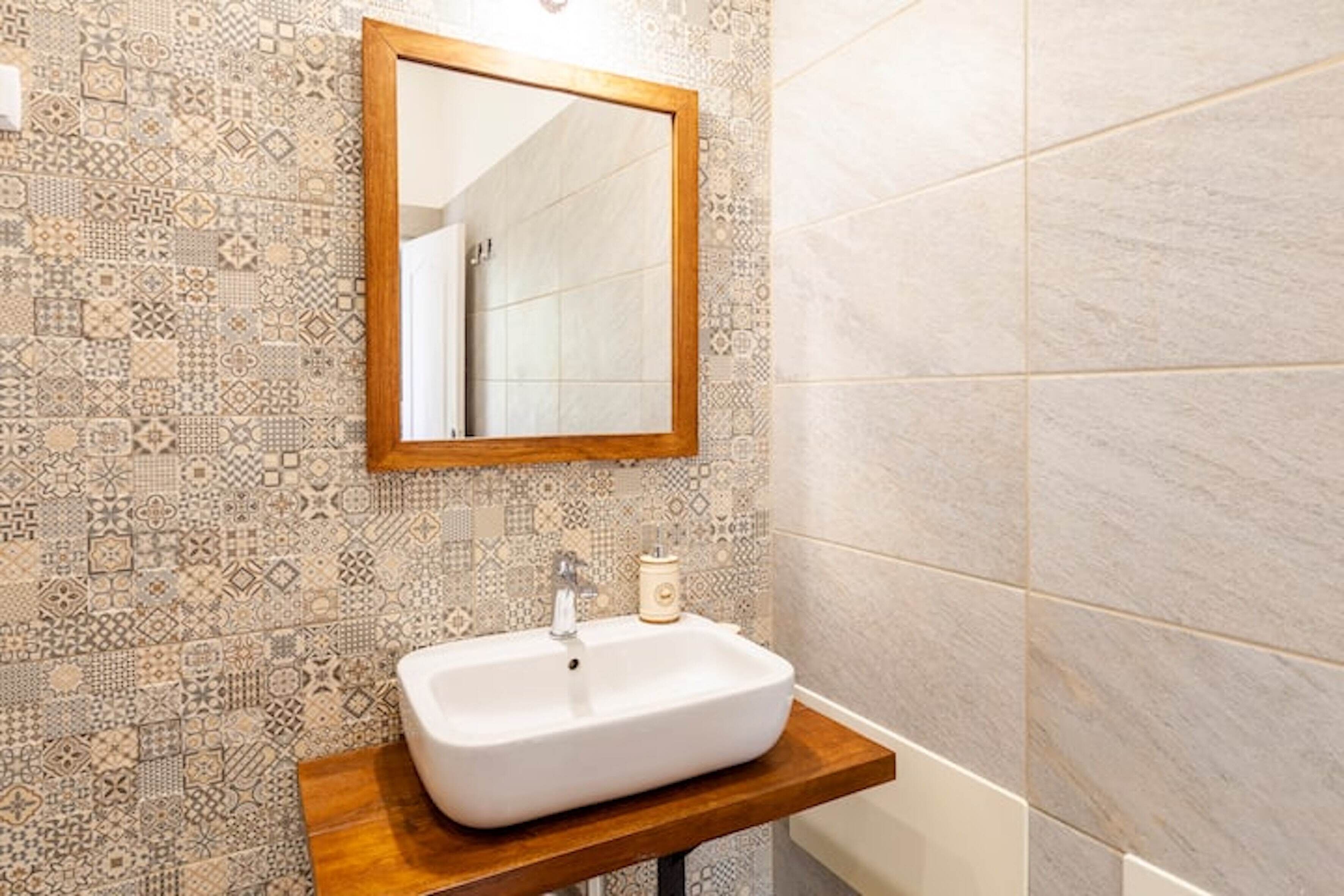 Casa de banho com painel de azulejos estampados, toucador de madeira com lavatório branco e espelho acima.