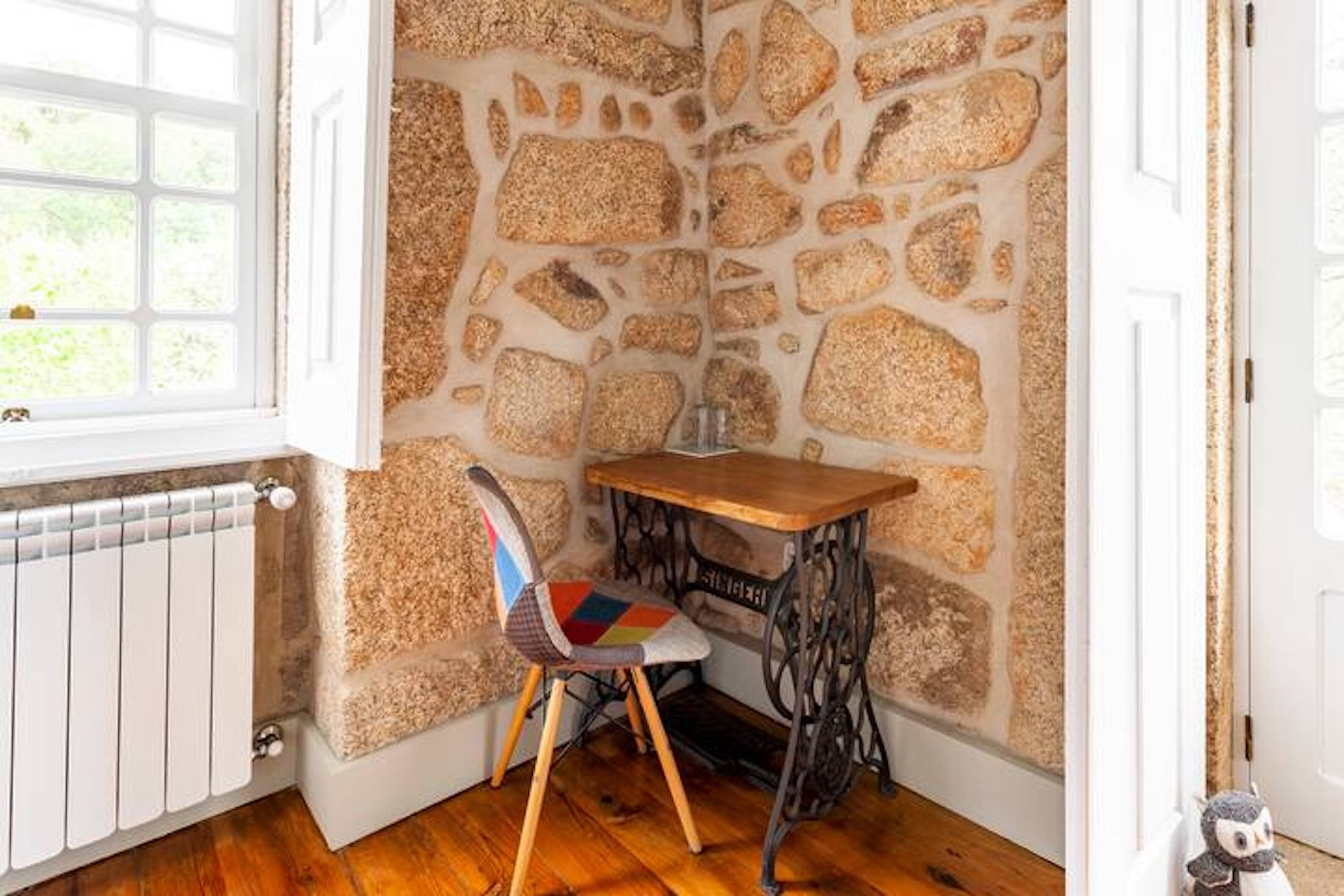 A corner room with a stone wall, a wooden table, a colorful chair, and a radiator.