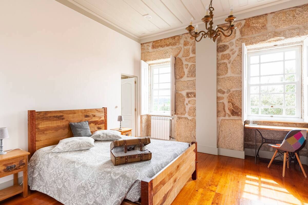 A bedroom with a wooden bed, stone walls, a desk, and a chair by a window.
