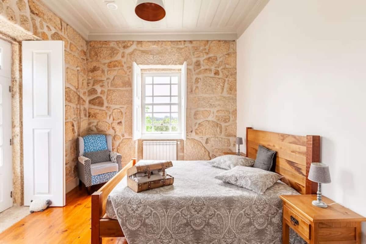 A bedroom with a stone wall, a wooden bed, a chair, and a window.