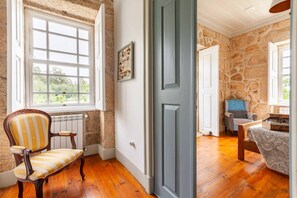 A room with a wooden floor, a stone wall, a blue door, a window with white frames, and a chair with striped upholstery. - Casa do Cabeço (Tondela)