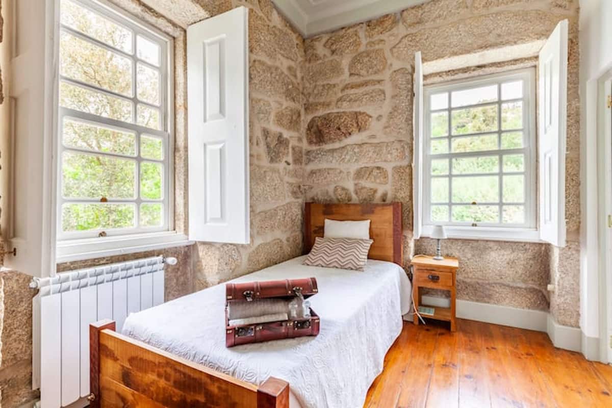 A bedroom with a stone wall, a wooden bed, two windows with white shutters, and a small bedside table.