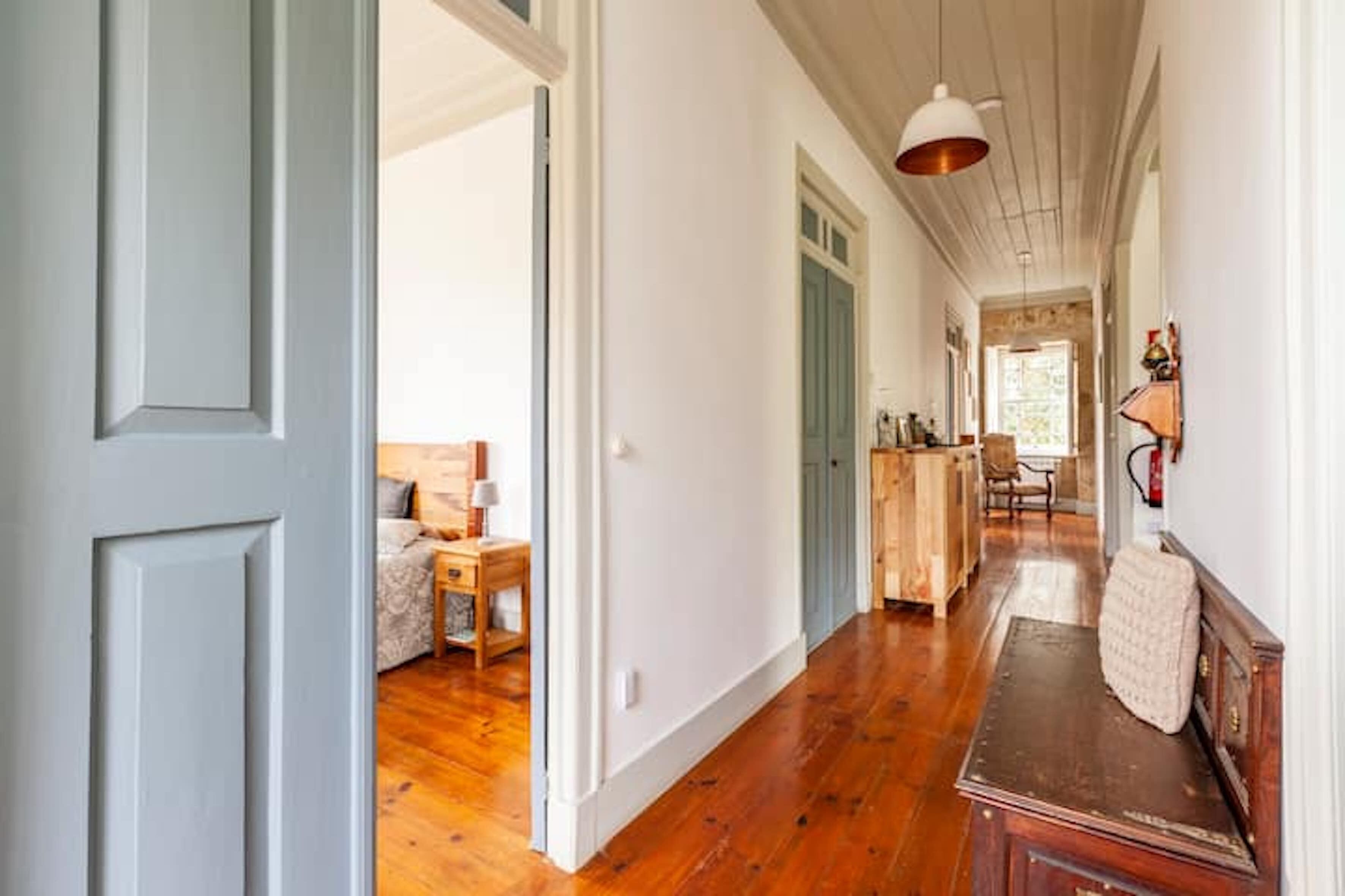 A hallway with wooden flooring, a bench, and a door leading to a room with a bed and a nightstand.