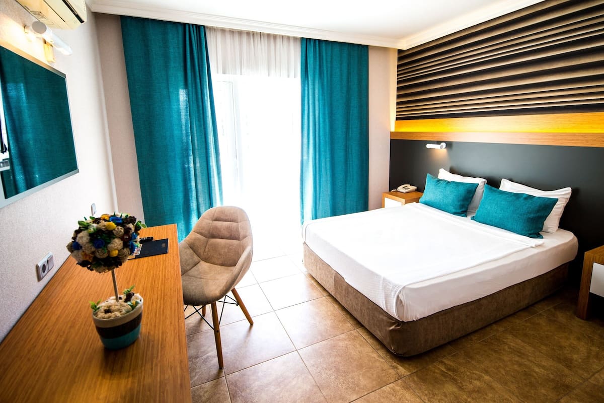 A hotel room with a bed, a desk, a chair, a potted plant, and a nightstand with a lamp.