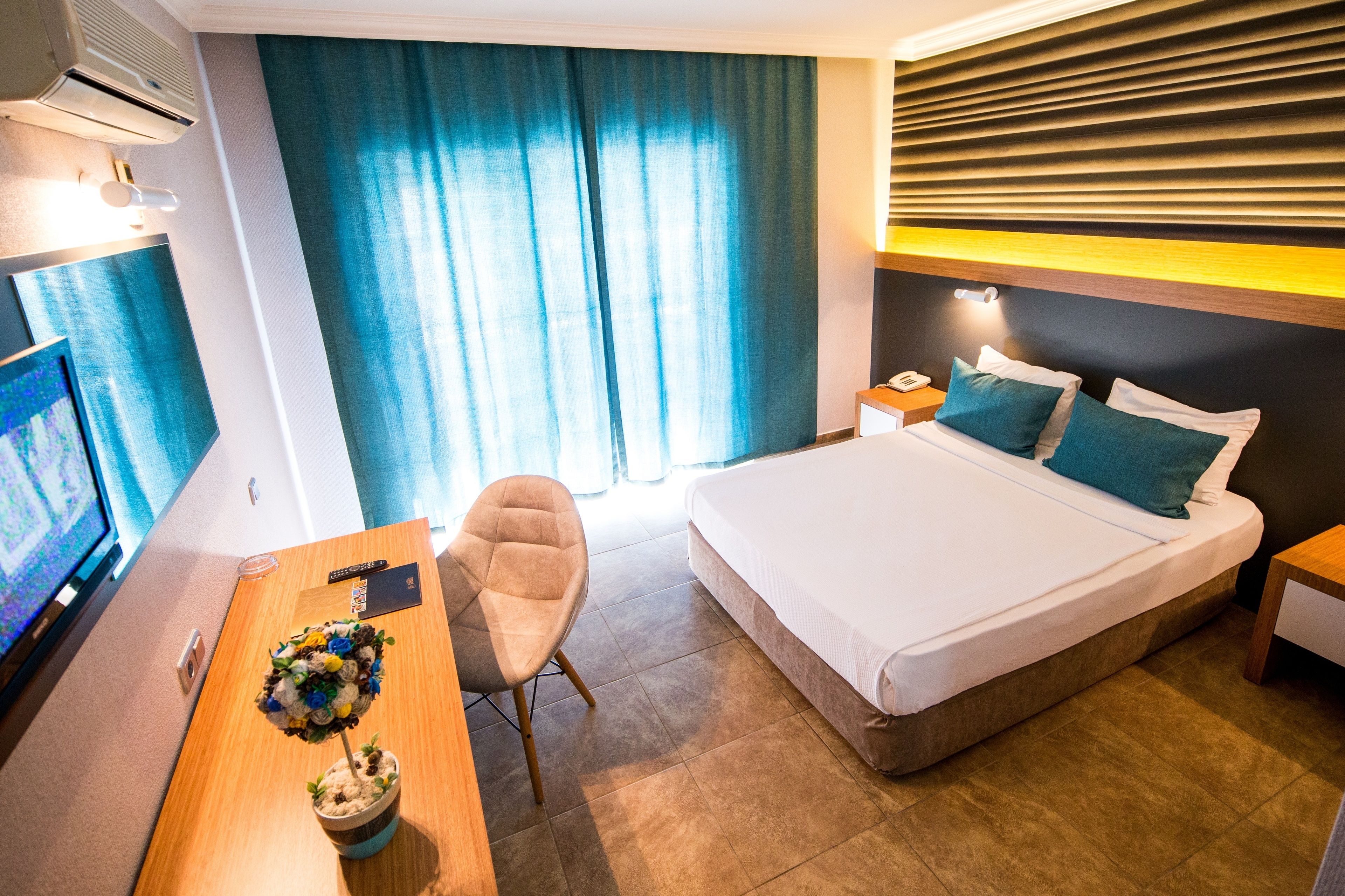 Quarto de hotel com cama, escrivaninha, cadeira, televisão e janela com cortinas.
