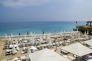 A beach with sun loungers and umbrellas, a pier with tables and a red sailboat, and the sea in the background. - Club Hotel Sunbel - All Inclusive (Kemer)