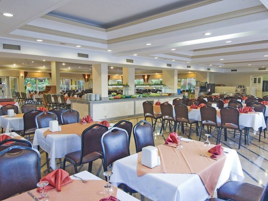 Un amplio salón de banquetes con numerosas mesas y sillas, una zona de buffet y una cocina al fondo.