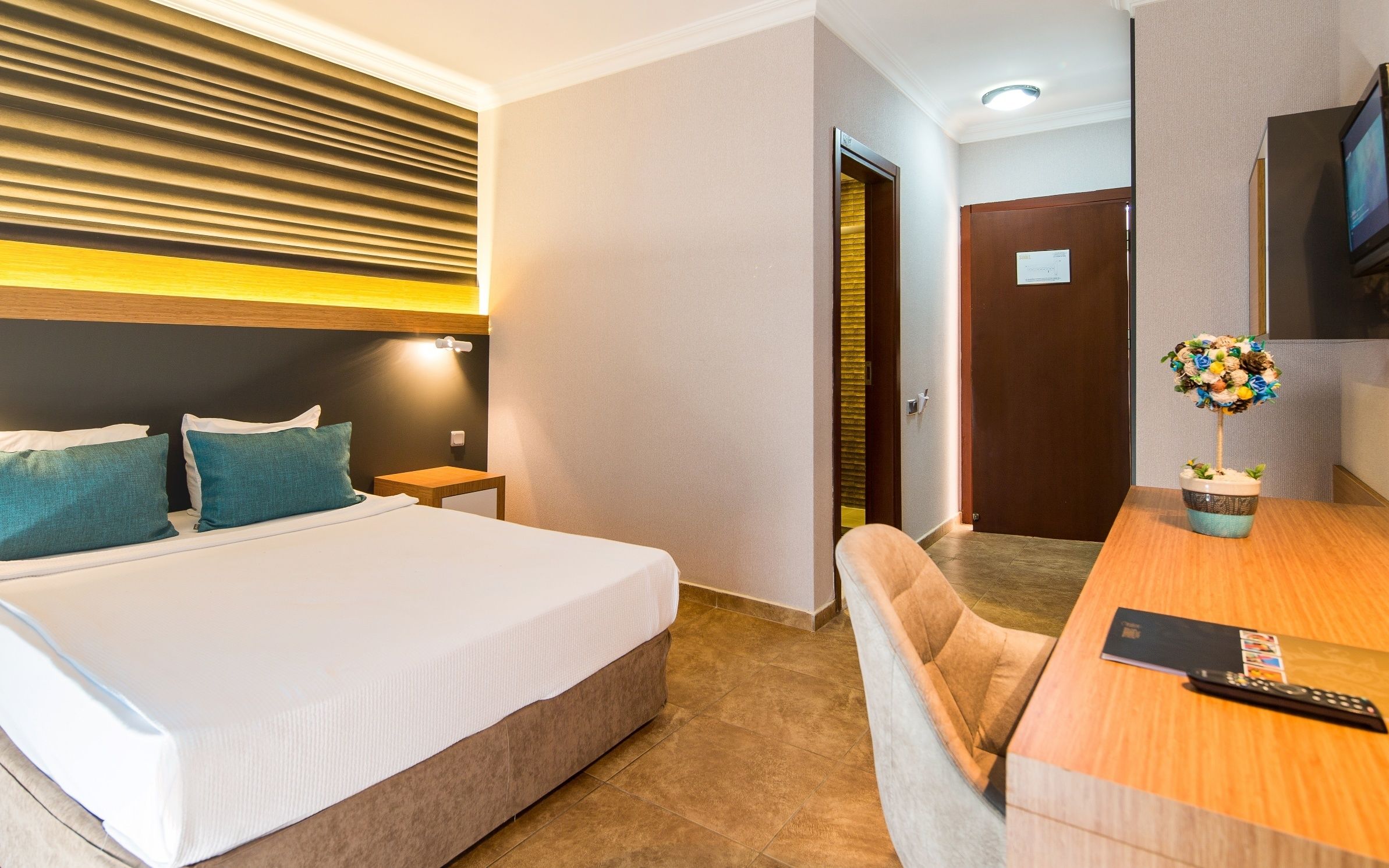 Habitación de hotel con cama, mesitas de noche, escritorio con silla, televisor y una planta decorativa.