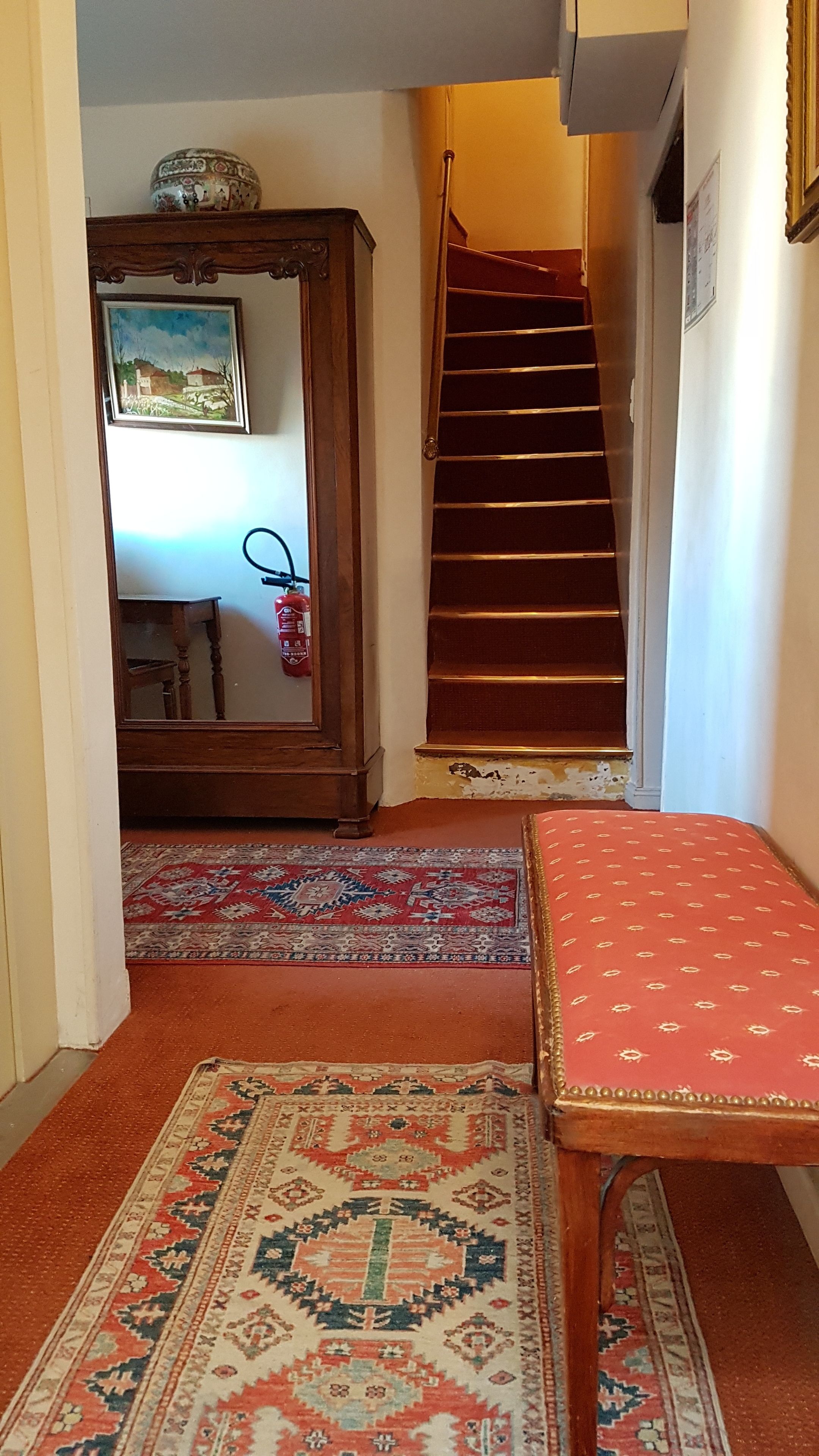 A hallway with a staircase, a wooden cabinet with a mirror, a red patterned rug, and a red cushioned bench.