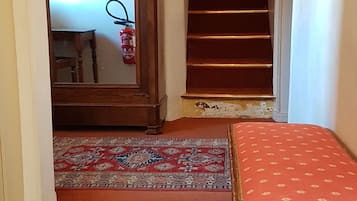 Un pasillo con una escalera, un mueble de madera con espejo, un tapete rojo estampado y un banco acolchado rojo.
