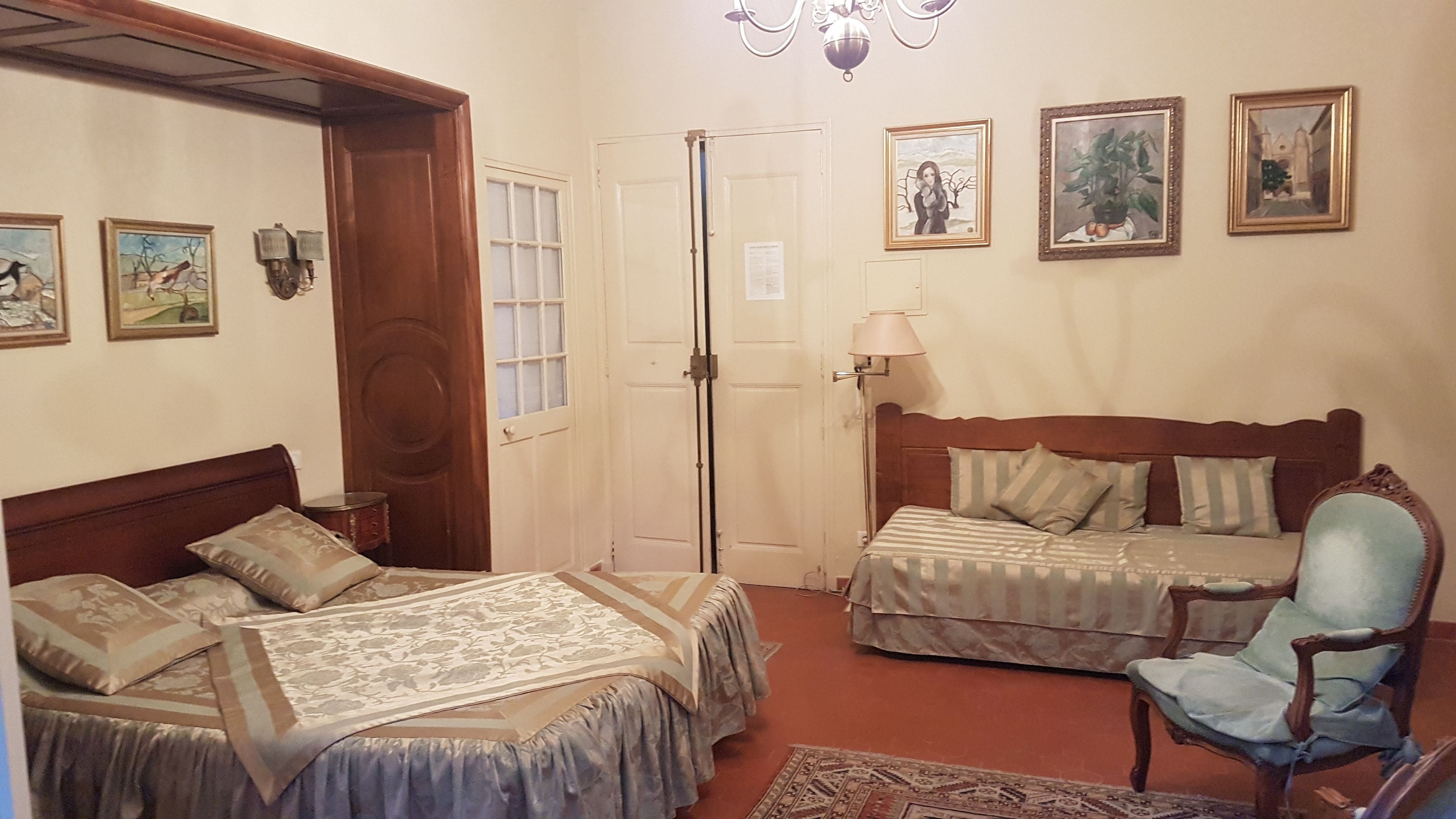 A bedroom with two beds, a chair, and a lamp. There are framed pictures on the wall.